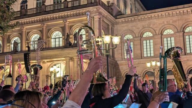 Eine Gruppe Musikerinnen und Musiker hebt ihre Blasinstrumente synchron vor einem historisch beleuchteten Theatergebäude bei Nacht. Die Szene strahlt Gemeinschaft und feierliche Stimmung im Freien aus.