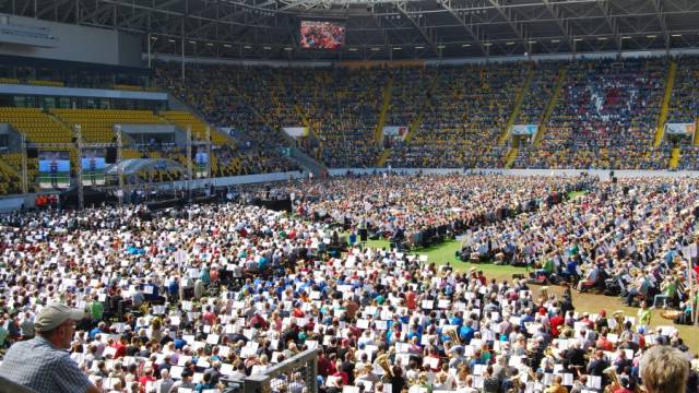 Tausende Menschen sitzen in einem großen Stadion eng beieinander und musizieren gemeinsam. Vor ihnen befinden sich Notenpulte und Blasinstrumente, während auf einer Bühne Bildschirme und Technik aufgebaut sind. Die voll besetzten Ränge und das sonnige Licht erzeugen eine festliche, beeindruckende Großveranstaltung.