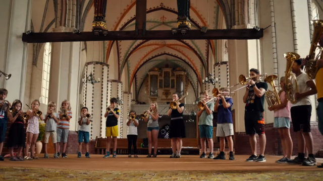 Eine Gruppe von Kindern und Jugendlichen steht in einer Reihe und spielt Blechblasinstrumente. Sie wirken konzentriert und musizieren gemeinsam. Der Auftritt findet in einer gro&szlig;en, hellen Kirche statt.