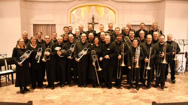 ChatGPT: Eine große Gruppe von Bläser*innen steht in einem Kirchenraum zusammen und hält verschiedene Blechblasinstrumente wie Trompeten, Hörner und Posaunen. Alle tragen dunkle Kleidung und lächeln in die Kamera. Hinter ihnen ist ein helles Altarbild mit goldenen Akzenten zu sehen.