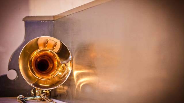 Der Schalltrichter einer goldglänzenden Posaune liegt auf einer glatten, hellen Oberfläche und spiegelt warmes Licht wider. Der Fokus liegt auf der runden Öffnung des Instruments. Der Hintergrund ist weich und unscharf gehalten.