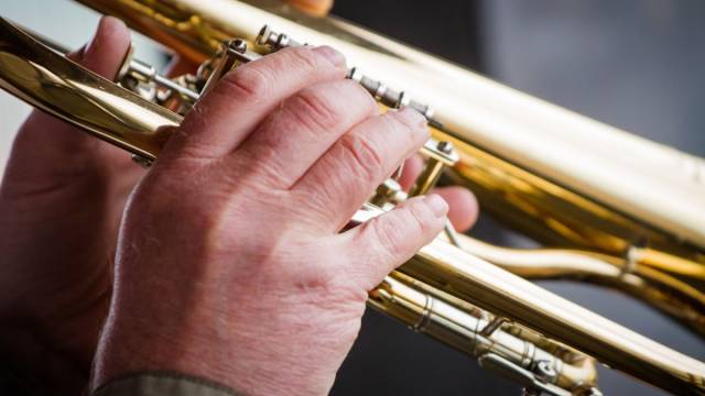 Nahaufnahme zweier Hände, die eine glänzende Trompete greifen und die Ventile bedienen. Die goldene Oberfläche des Instruments reflektiert das Licht. Der Hintergrund ist unscharf und lenkt den Fokus auf das Musizieren.