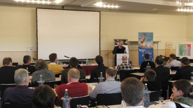 In einem Konferenzraum sitzen zahlreiche Teilnehmende an Tischen und hören einem Vortragenden am Rednerpult zu. Hinter ihm steht eine große Leinwand sowie mehrere Informationsbanner. Wasserflaschen, Unterlagen und Gläser liegen auf den Tischen der Zuhörenden.