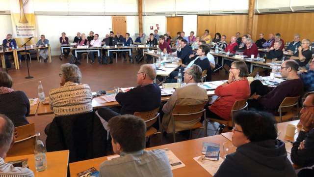 Viele Menschen sitzen in einem großen Tagungsraum an Tischen und hören einer Sitzung oder Präsentation zu. Auf den Tischen liegen Unterlagen, Getränke und Arbeitsmaterialien. Im Hintergrund hängt ein Banner des Evangelischen Posaunendienstes in Deutschland.