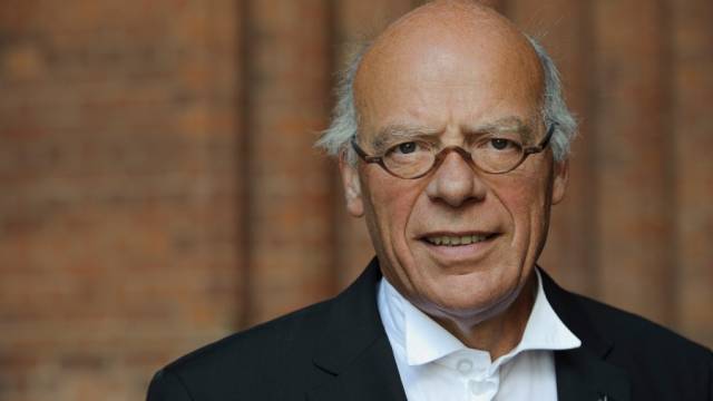 Eine Person mit rundlicher Brille und lichtem, grauem Haar blickt direkt in die Kamera. Sie trägt ein weißes Hemd und ein dunkles Jackett. Der Hintergrund besteht aus unscharfem Mauerwerk in warmen Farbtönen.