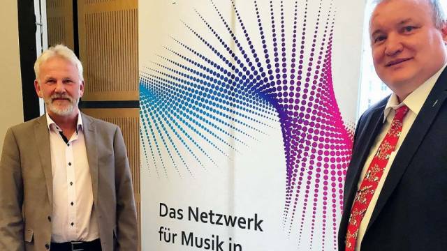 Zwei Männer in Anzugkleidung stehen vor einem Banner mit farbigem Punktmuster und der Aufschrift „Das Netzwerk für Musik in Deutschland“. Beide lächeln in die Kamera. Die Szene wirkt wie ein Treffen oder eine Veranstaltung in einem Innenraum.