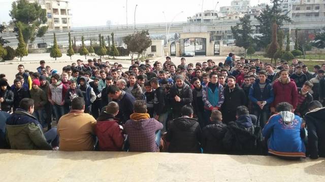 Eine große Gruppe junger Menschen steht und sitzt dicht beieinander auf einem Platz vor Gebäuden. Einige hören aufmerksam zu, andere sprechen miteinander. Im Hintergrund sind Straßen, Bäume und städtische Architektur zu sehen.