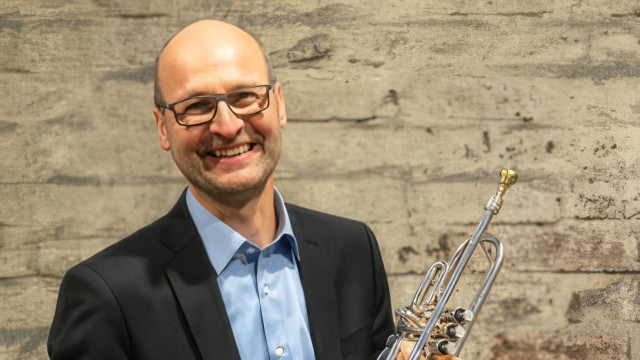 Porträt eines lächelnden, glatzköpfigen Mannes mit Brille im dunklen Sakko und hellblauem Hemd; er hält eine Trompete vor einer grauen, rauen Wand