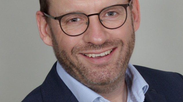 Porträt eines lächelnden Mannes mit runder Brille und kurzem Bart; er trägt hellblaues Hemd und dunkles Sakko vor neutralem, hellem Hintergrund