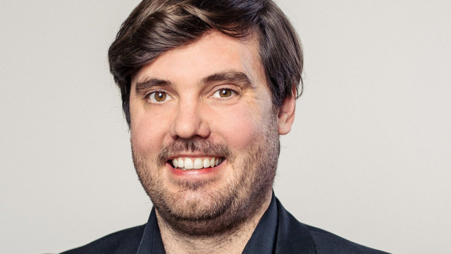 Porträt eines lächelnden Mannes mit kurzem grauem Haar und Brille in dunklem Sakko; Hintergrund hell und unscharf