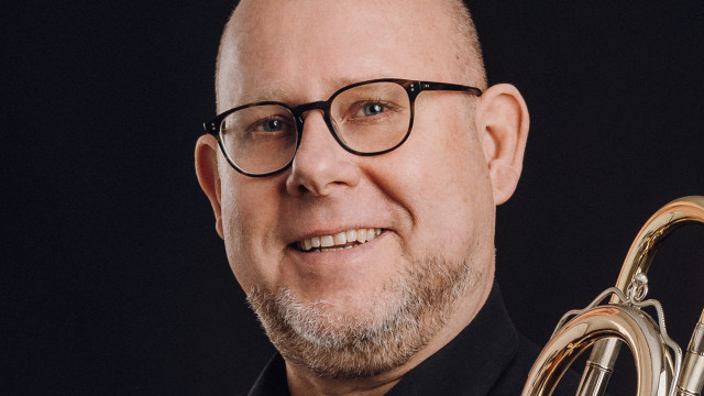 Frank Möwes Porträt eines lächelnden Mannes mit Brille, schwarzem Hemd und einem Blechblasinstrument vor dunklem Hintergrund