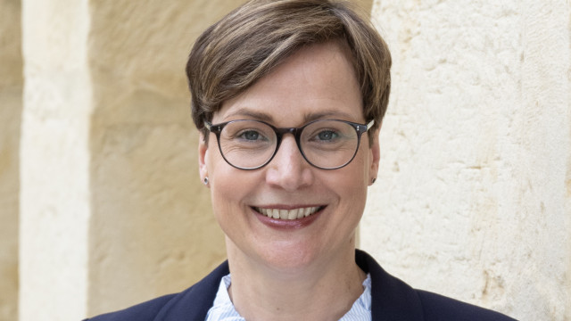 Marianne Gorka Porträt einer lächelnden Frau mit Brille, kurzem Haar und Blazer vor hellem Stein-Hintergrund