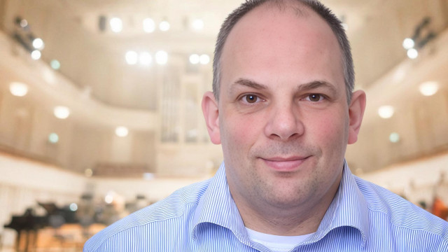 Dr. Frank Beekmann Porträt eines lächelnden Mannes im karierten Hemd vor einer holzfarbenen Wand