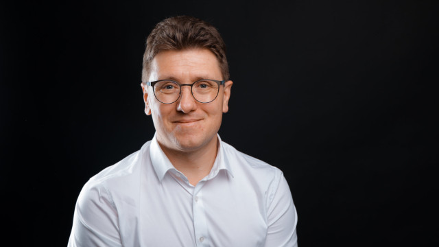 Sebastian Haase Mann mit Brille und weißem Hemd vor schwarzem Hintergrund, lächelnd.